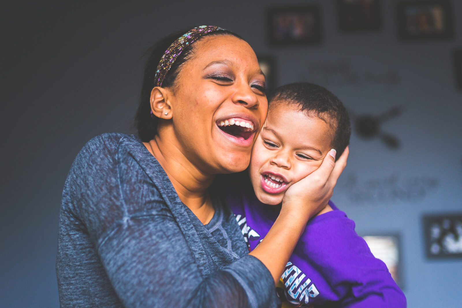 A woman with a floral headband and a gray top shares a genuine moment of laughter with a young boy in a purple shirt. The woman is smiling widely with her mouth open in a joyful laugh, her eyes slightly closed. She gently holds the boy's head against her cheek, both displaying expressions of happiness and affection. They are indoors, with a blurred background featuring picture frames on the wall.